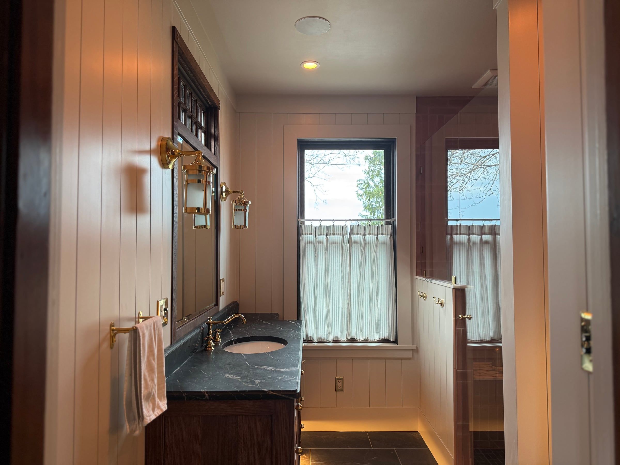 The bathroom after, from the doorway: white paneled walls, the oak vanity and mirror, and the window with a café curtain