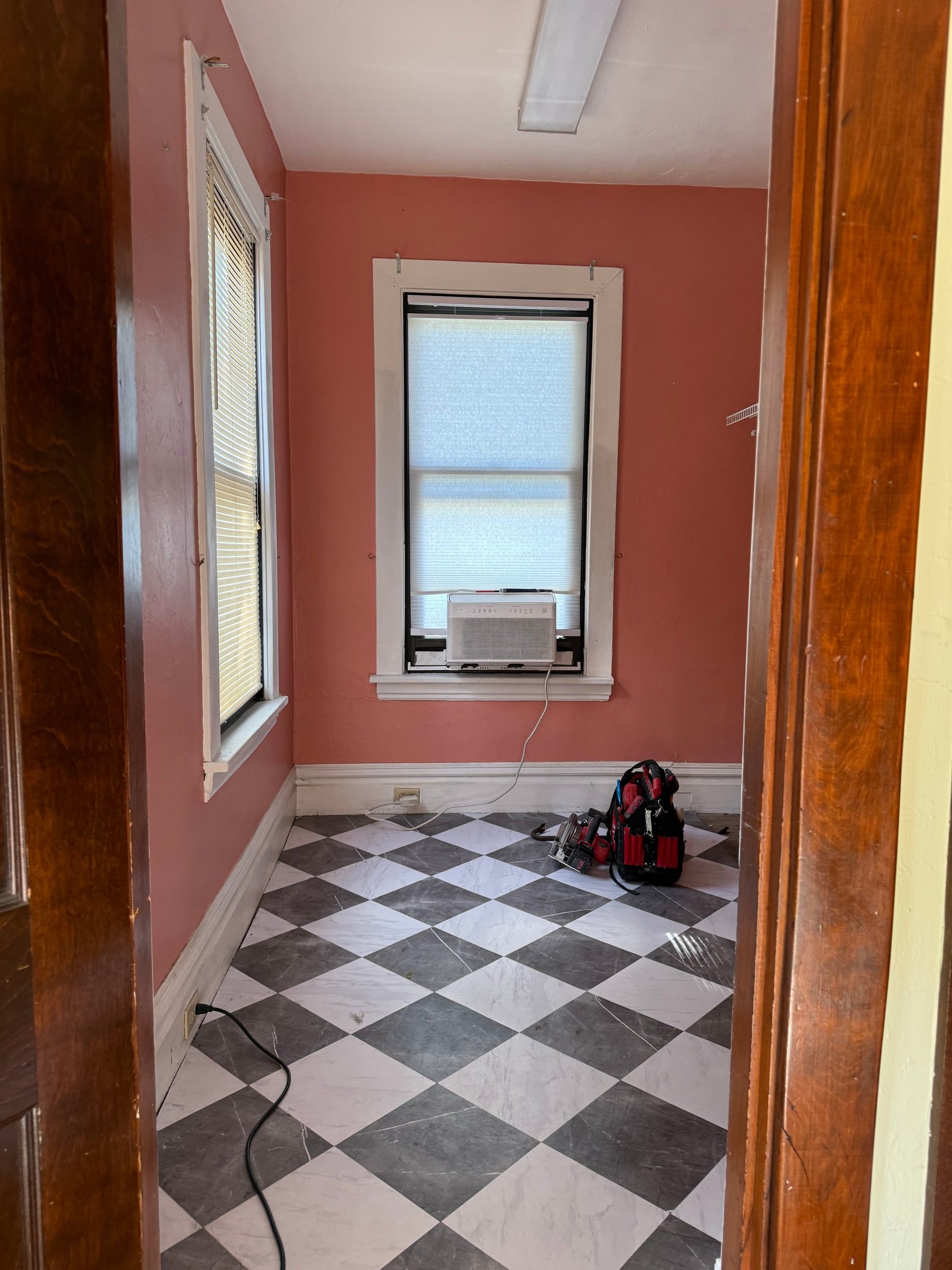 The bathroom before, from the doorway: pink walls, a black and white checkered floor, and a window air conditioner
