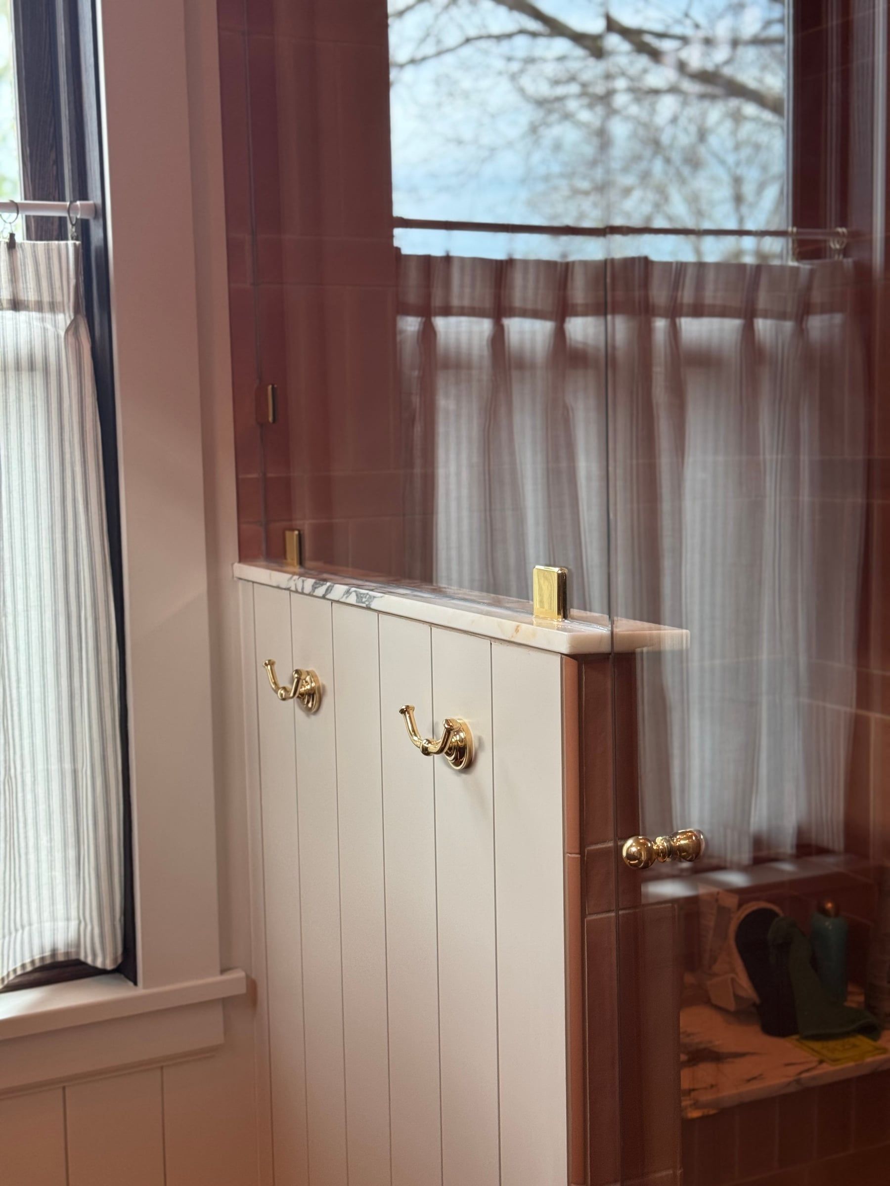 A paneled half wall with brass robe hooks, capped in marble with a glass panel into the shower, beside a window with café curtains