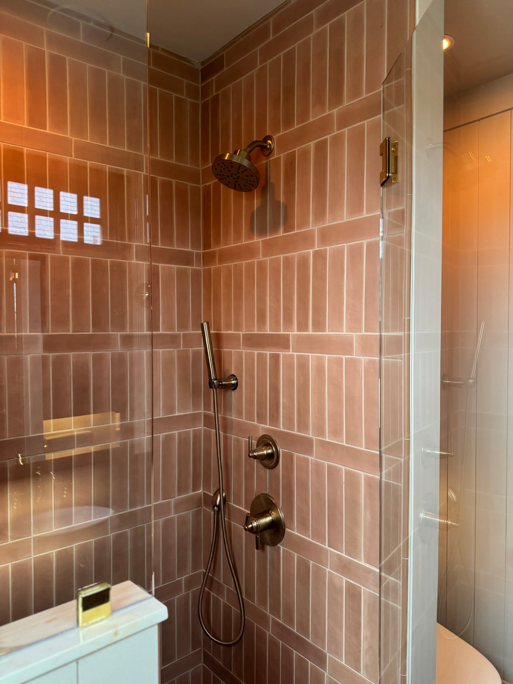 The shower: vertical terracotta-pink tile, brass fixtures, and a glass enclosure