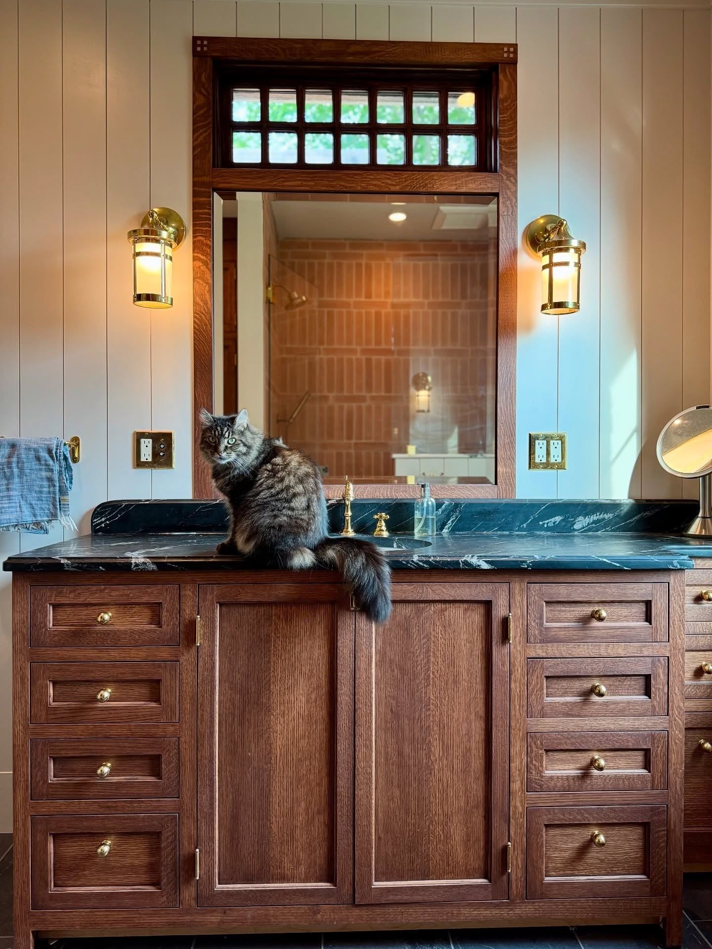 A quarter-sawn oak vanity with inset drawers and doors and brass knobs, under a mirror topped with a transom of small glass panes, between two brass sconces; a cat sits on the black stone counter
