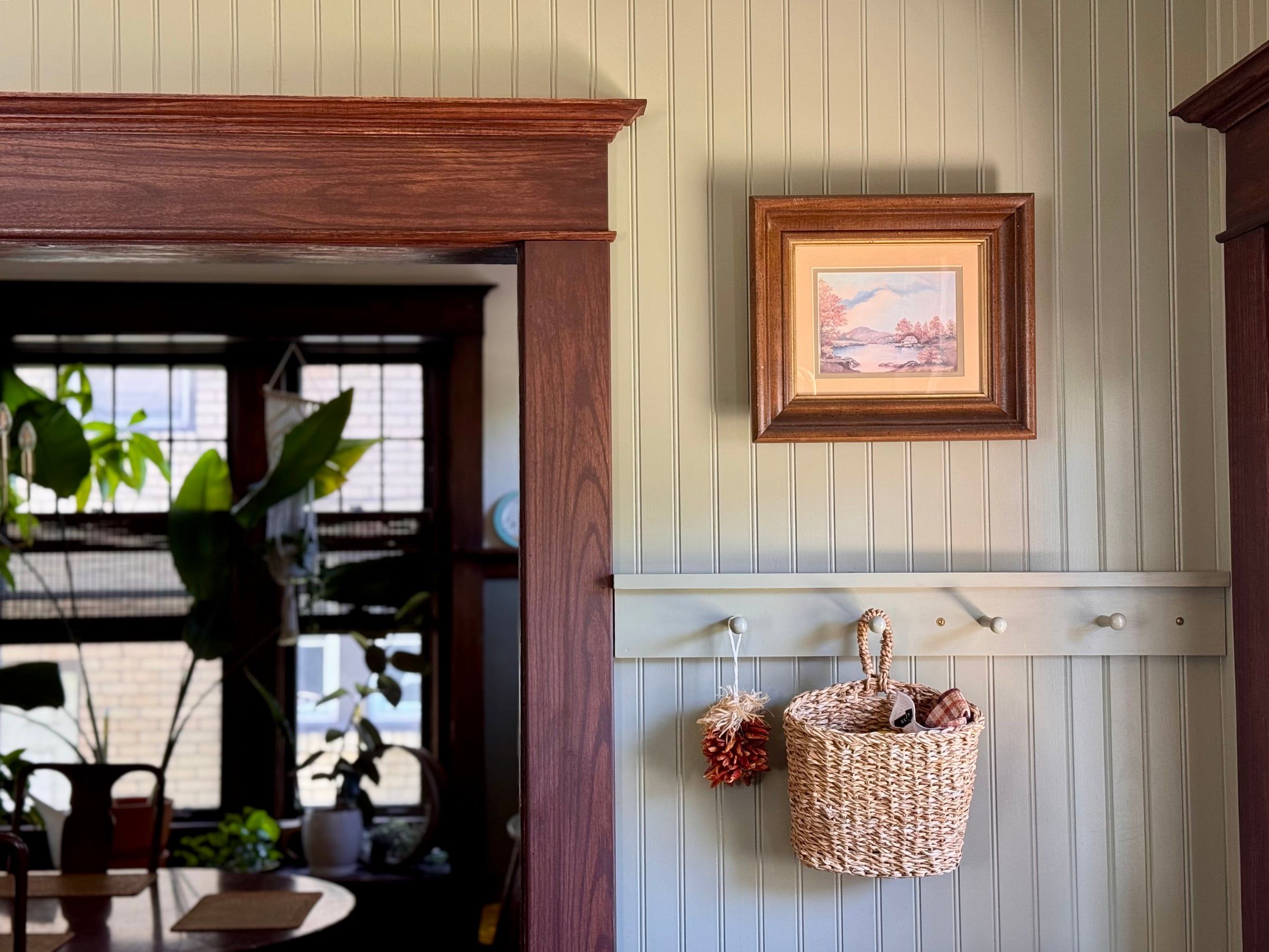 A dark-stained reproduction doorway casing with a cornice head, beside a beadboard wall with a framed painting, a peg rail, and a hanging basket