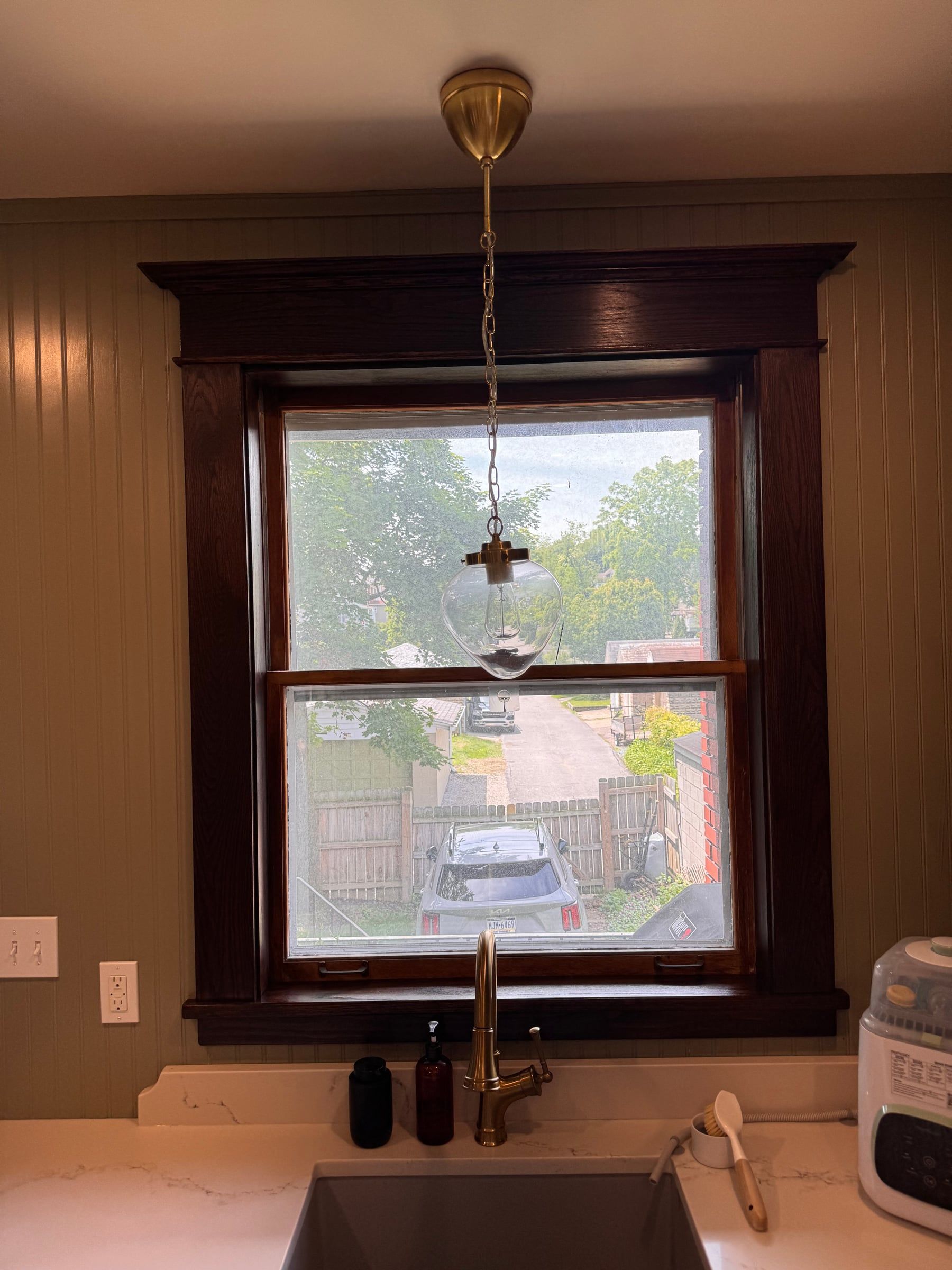 The kitchen window over the sink, framed in dark-stained reproduction Craftsman casing with a cornice head, against beadboard walls