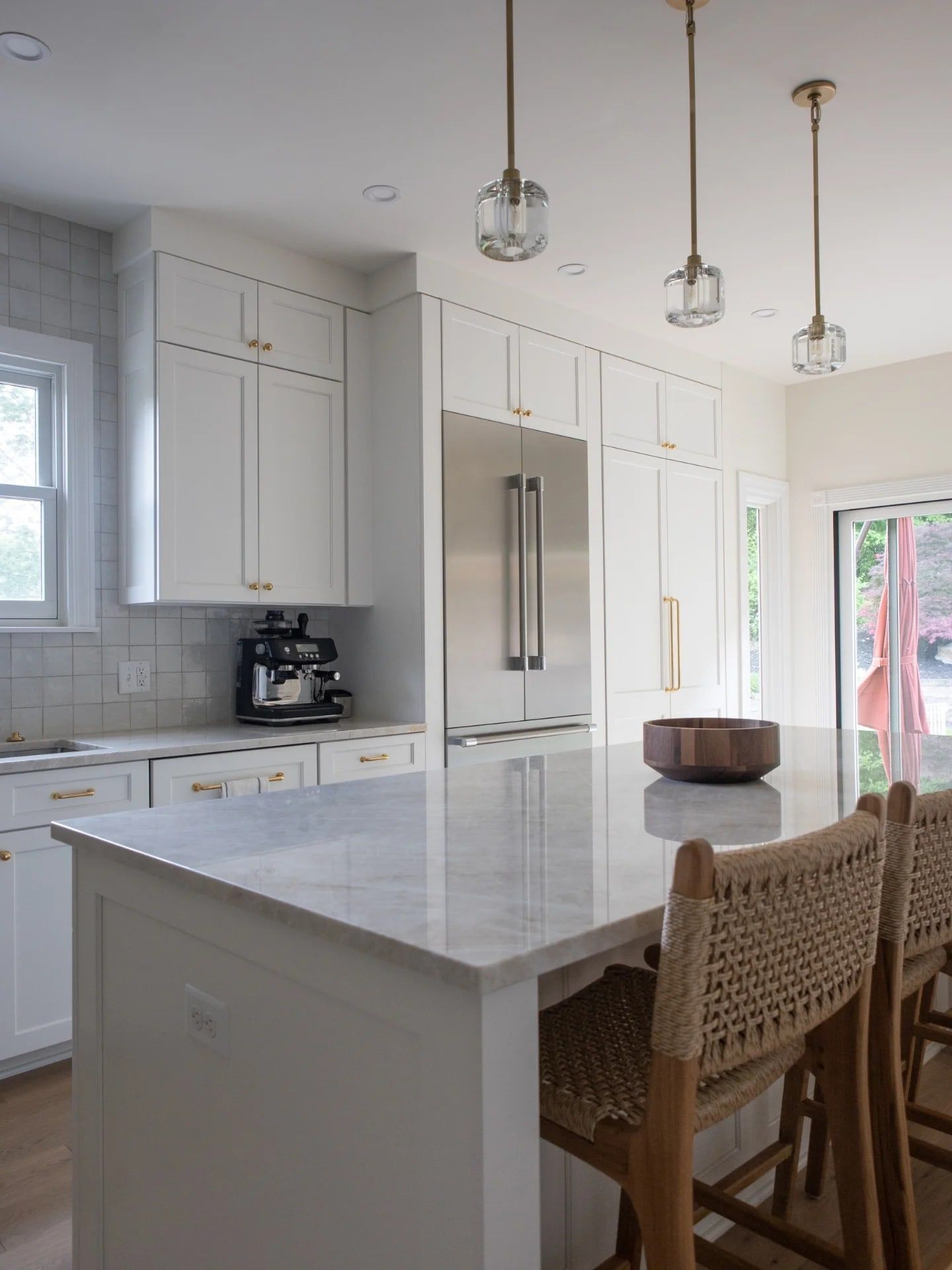 The kitchen after: an expanded island with a pale stone top, glass pendants, light painted cabinets, and a built-in refrigerator and pantry