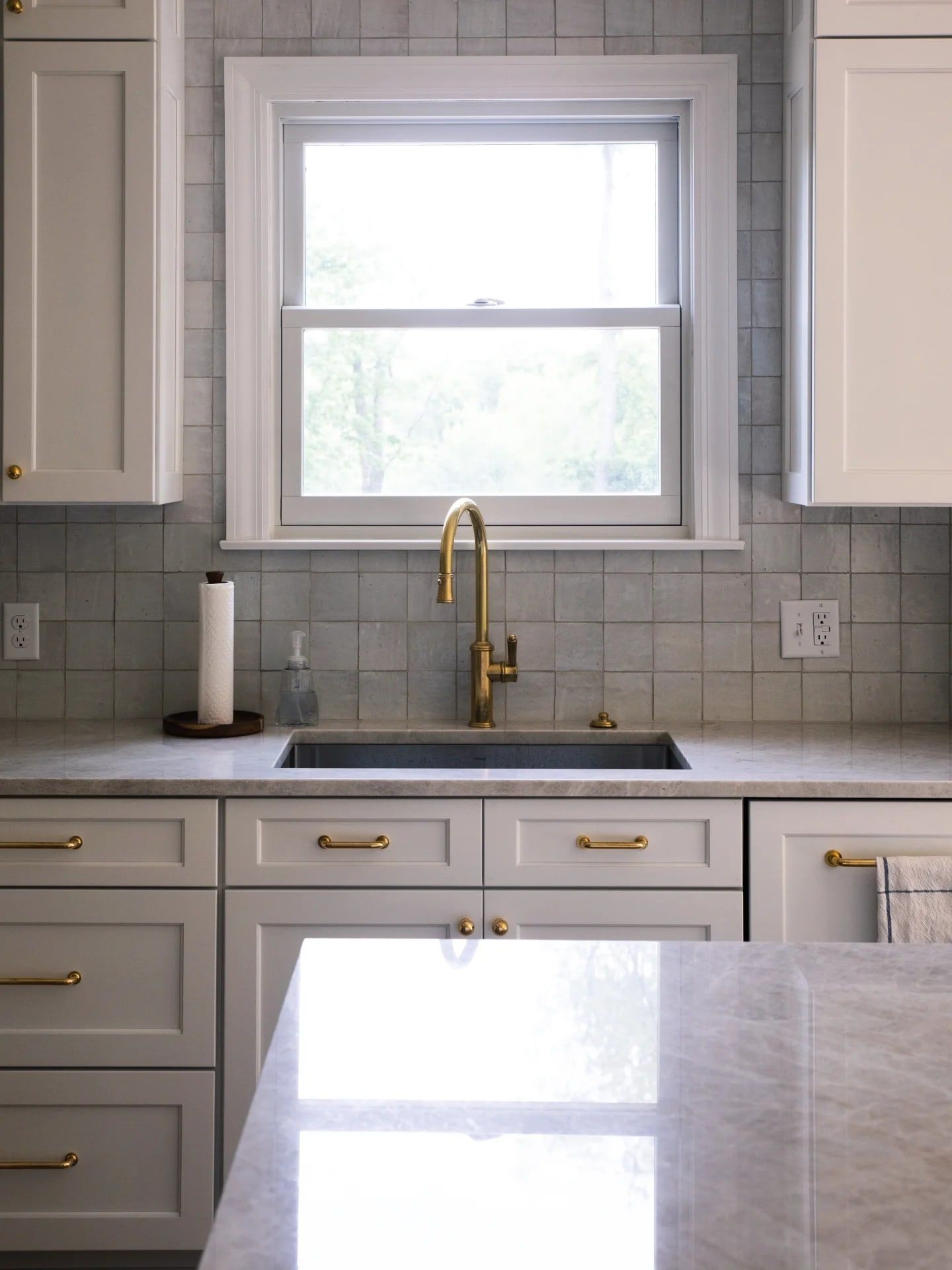 An unlacquered brass faucet at the sink under the window, with handmade zellige tile and brass cabinet hardware