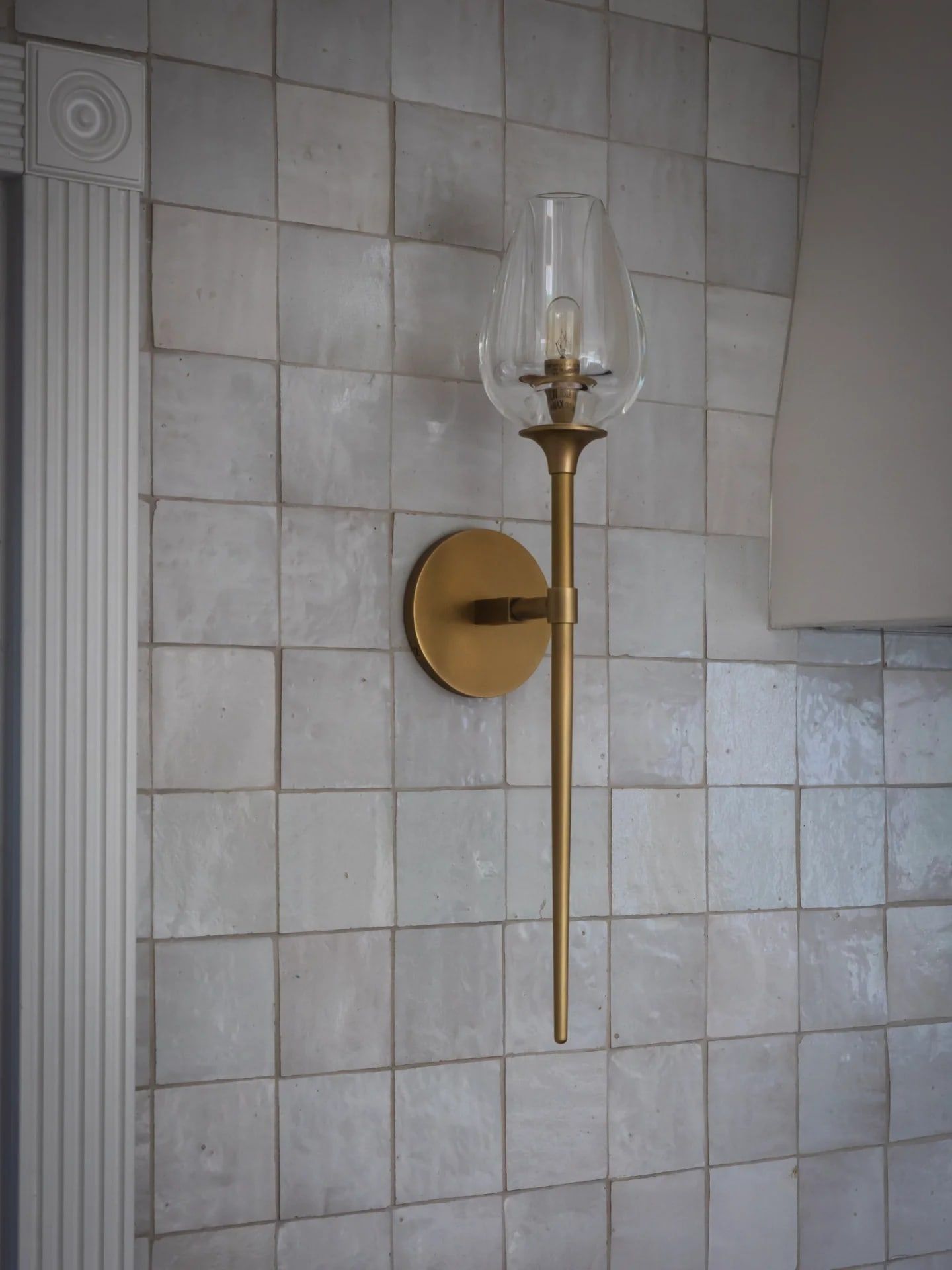 A tall brass sconce on a wall of handmade zellige tile, beside a fluted pilaster