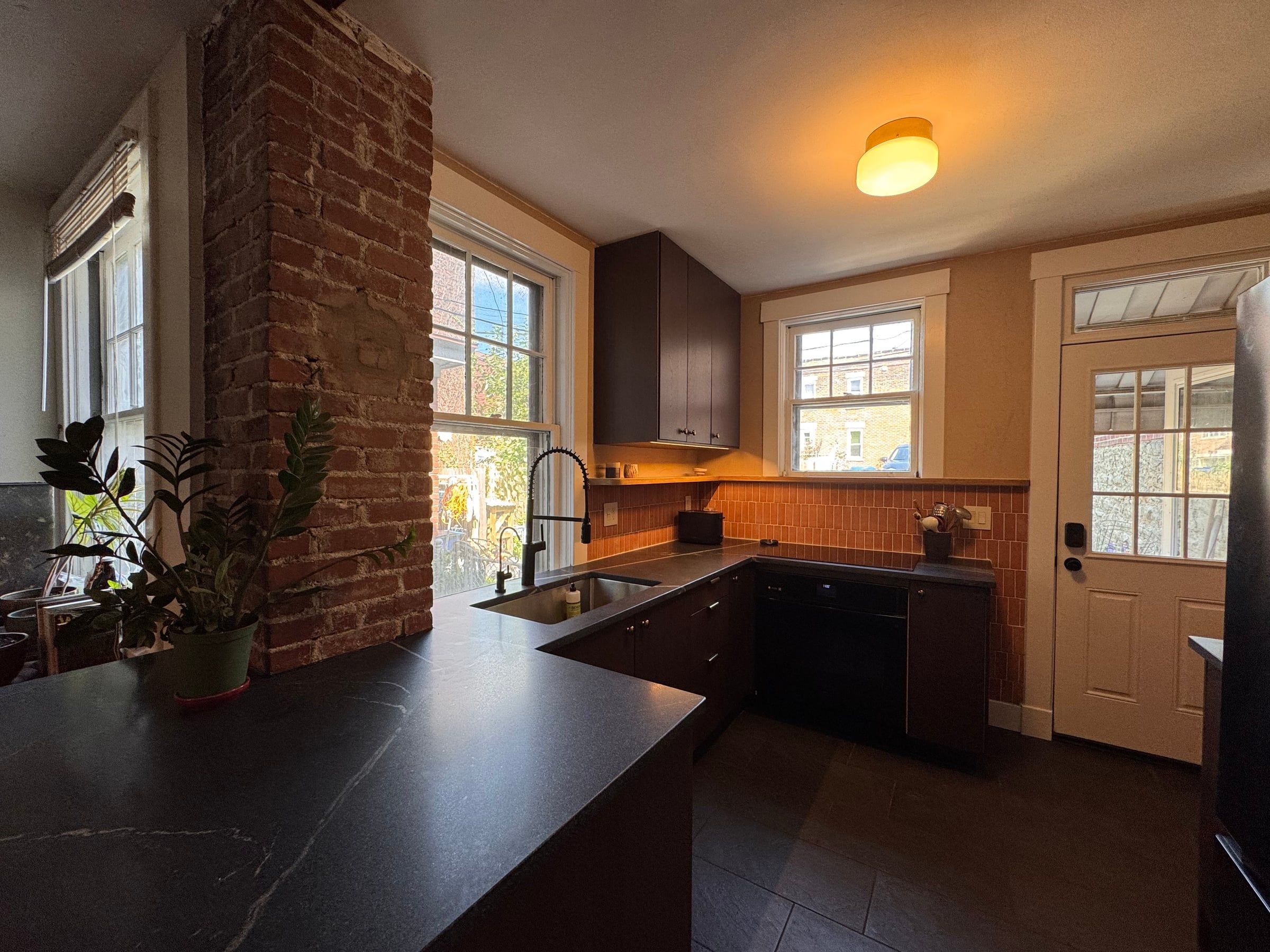 The finished kitchen from the corner: an exposed brick chimney, dark stained oak cabinets, a dark granite counter, handmade terracotta tile, and windows on two walls