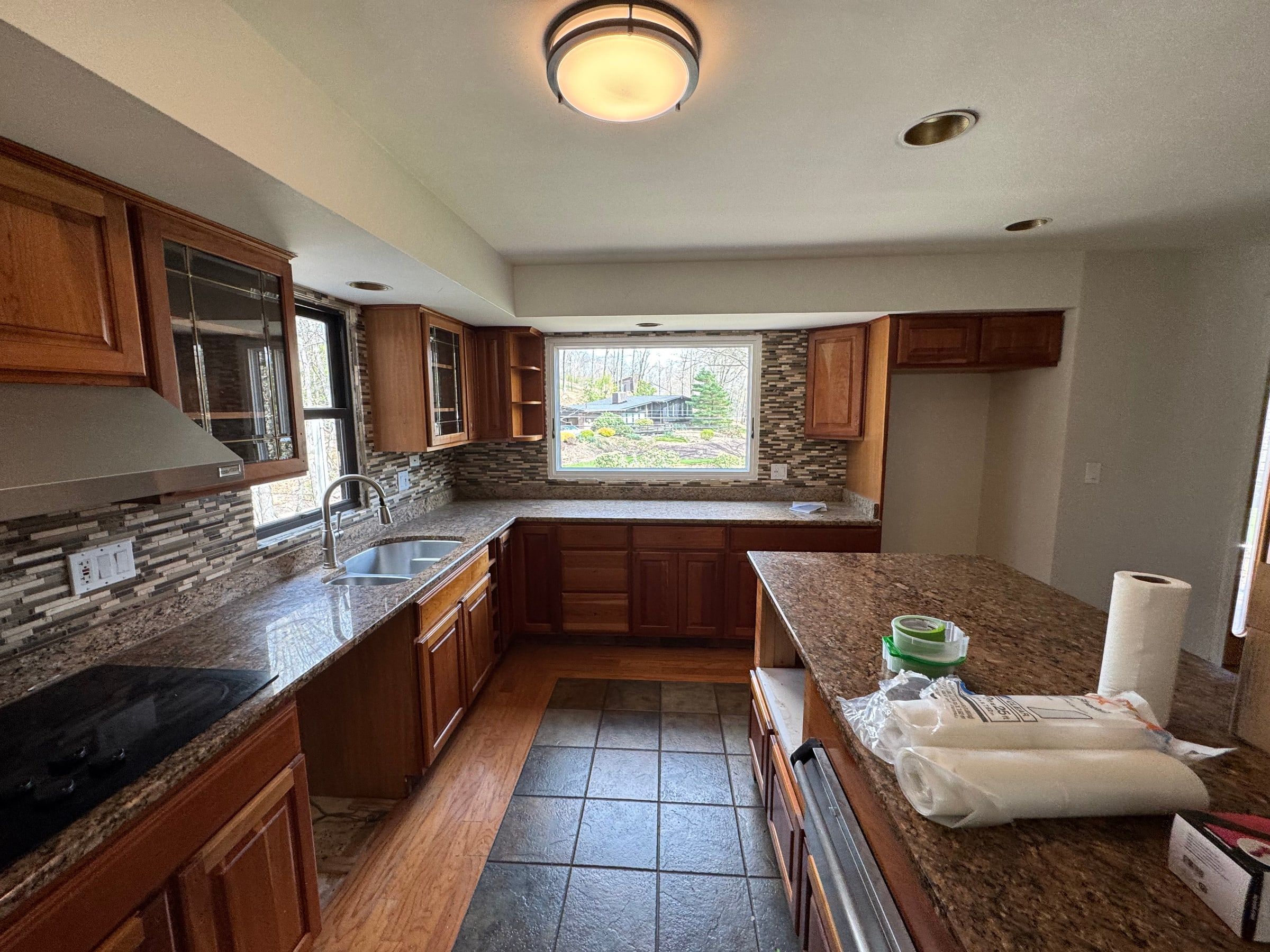The kitchen before: reddish wood cabinets, speckled granite, a glass mosaic backsplash, and a window looking out to the trees