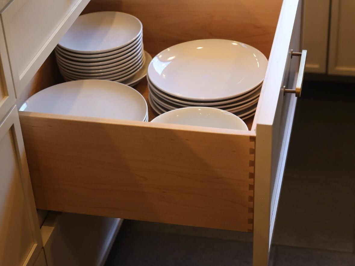 A deep drawer pulled open, its dovetailed wood box full of white plates, behind a thick inset drawer front with a brass pull