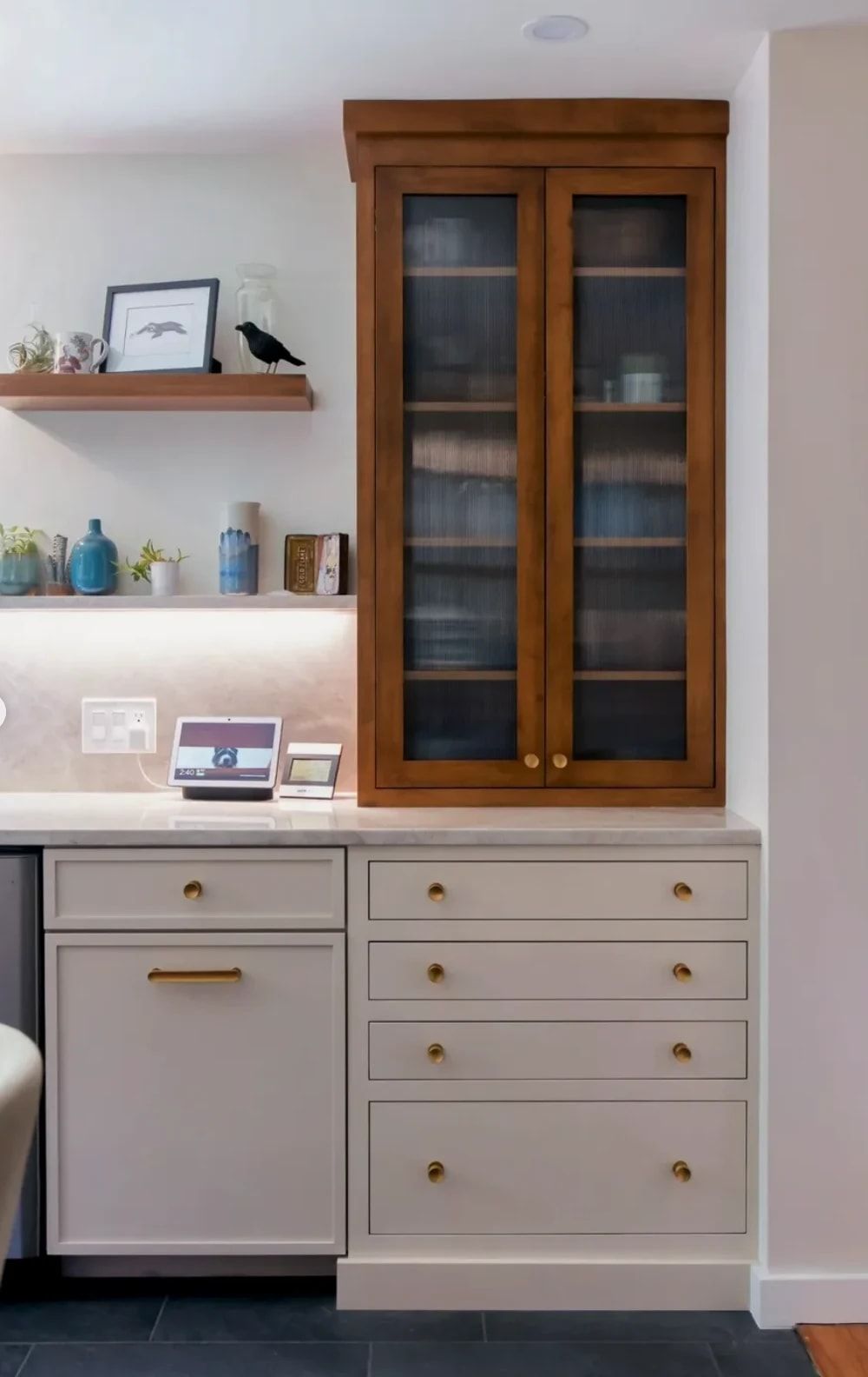 A warm wood cabinet with reeded glass doors sitting on the stone counter, above a stack of pale inset drawers, with floating shelves beside it