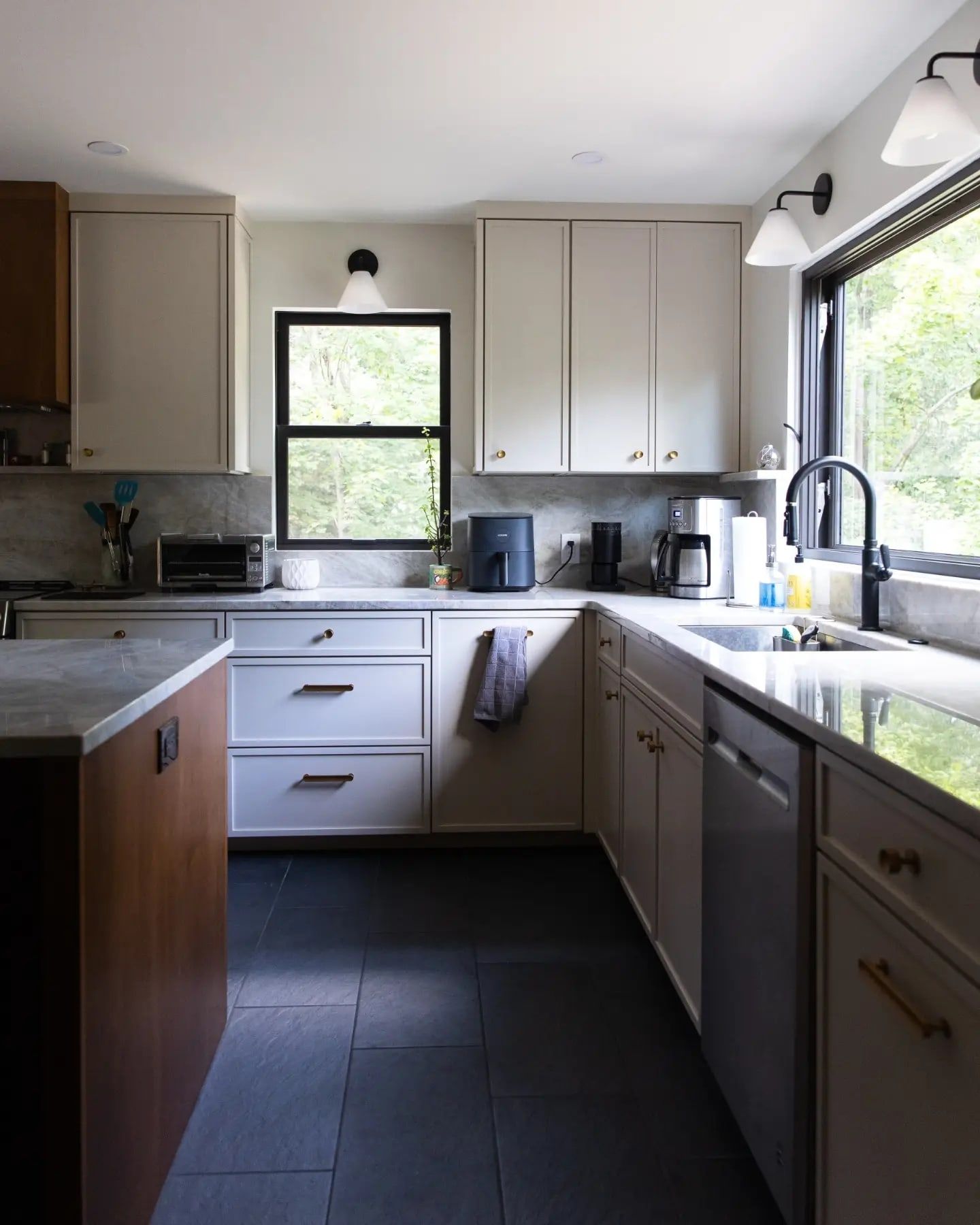 The finished kitchen: pale inset and overlay cabinets, a warm wood island with a pale stone top, black-framed windows, and dark slate floors