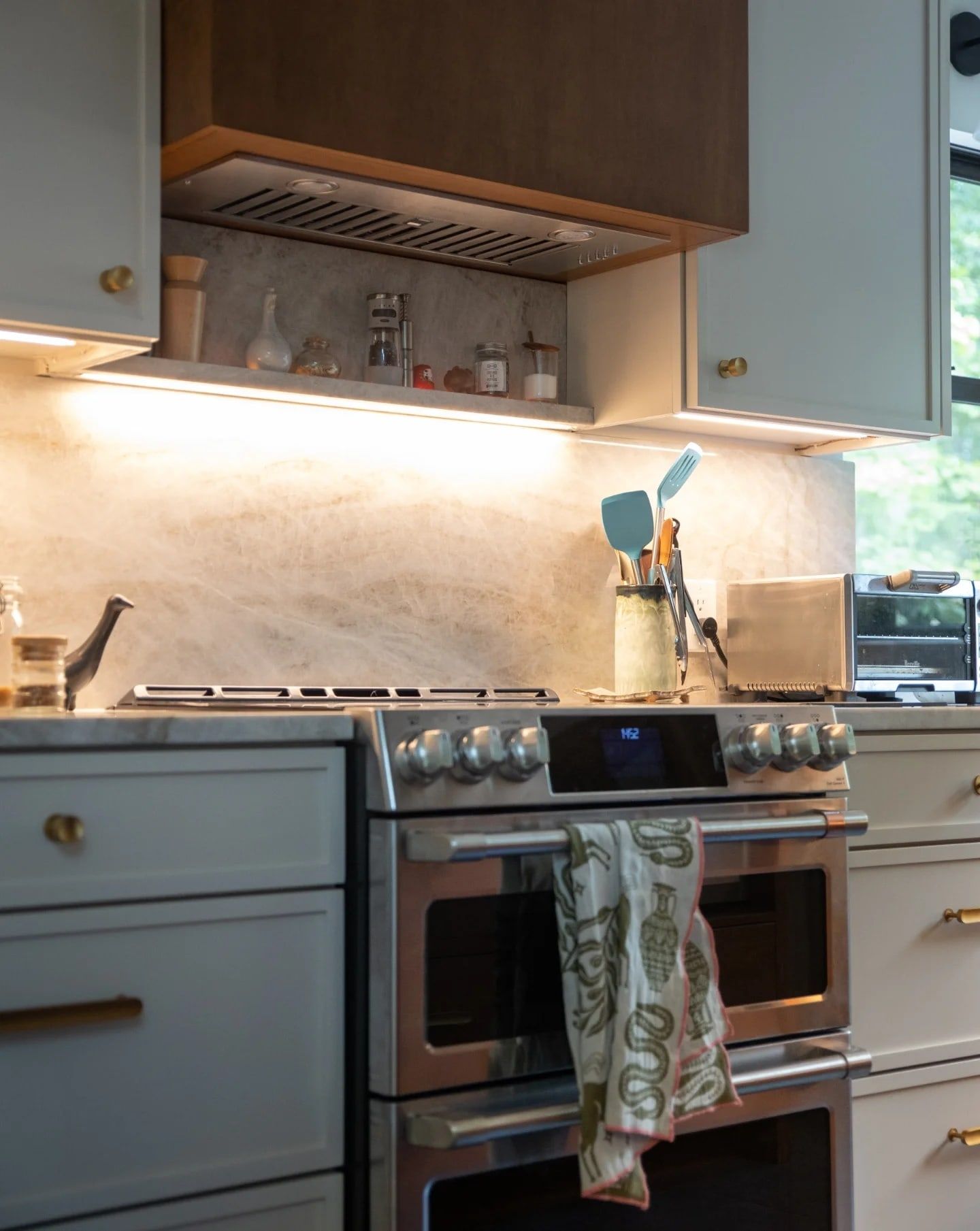 The range under a warm wood hood, with a stone slab backsplash and a lit stone ledge for spices