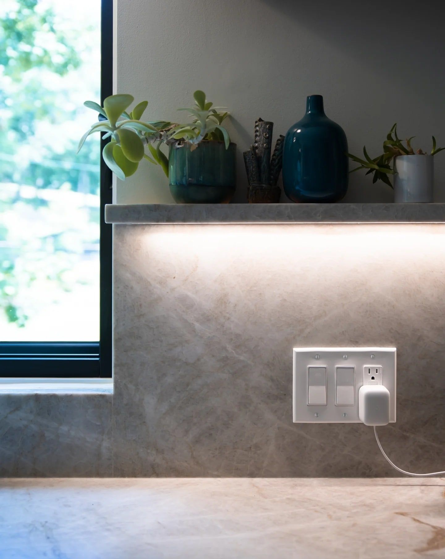 A stone ledge holding the homeowner’s pottery and plants, lit from a hidden channel beneath it that washes the stone backsplash