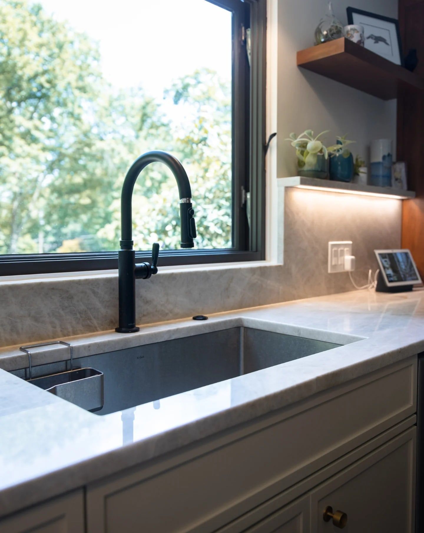 A black faucet and undermount sink in pale veined stone, below a black-framed window and a wood shelf
